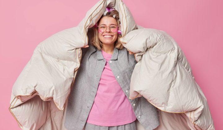 positive-cheerful-young-woman-wrapped-duvet-has-happy-expression-after-healthy-sleep-wakes-up-good-mood-wears-comfortable-pajama-makes-hairstyle-with-rollers-poses-against-pink-background