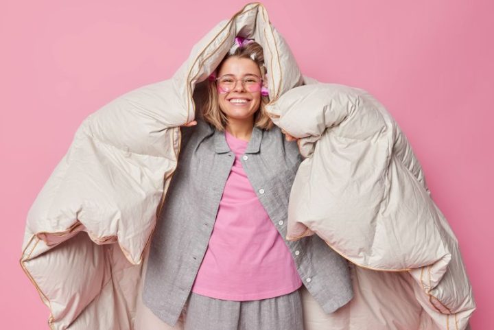 positive-cheerful-young-woman-wrapped-duvet-has-happy-expression-after-healthy-sleep-wakes-up-good-mood-wears-comfortable-pajama-makes-hairstyle-with-rollers-poses-against-pink-background
