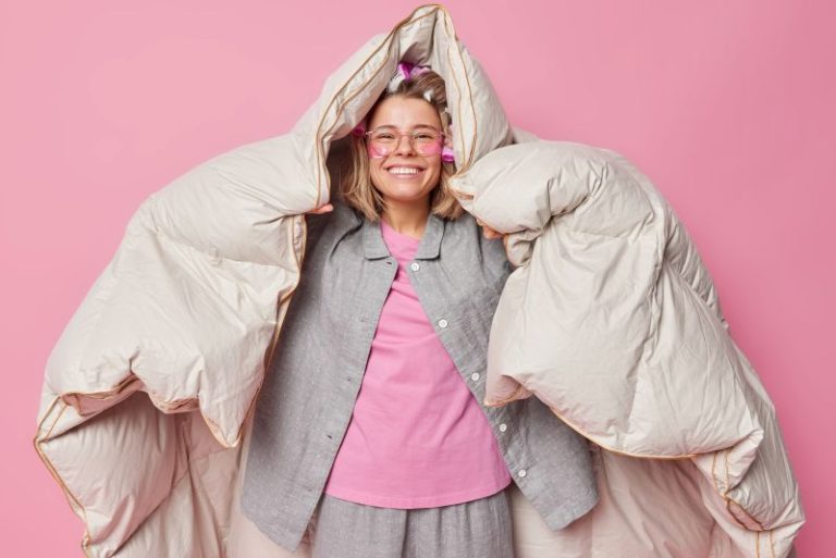 positive-cheerful-young-woman-wrapped-duvet-has-happy-expression-after-healthy-sleep-wakes-up-good-mood-wears-comfortable-pajama-makes-hairstyle-with-rollers-poses-against-pink-background