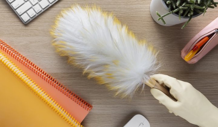 A hand in a glove holds a feather duster above a wooden desk with a keyboard, colorful notebooks, a potted cactus, a pencil case, and a wireless mouse.