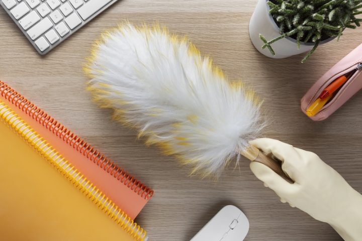 A hand in a glove holds a feather duster above a wooden desk with a keyboard, colorful notebooks, a potted cactus, a pencil case, and a wireless mouse.