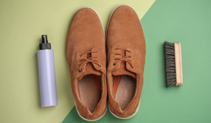 Brown suede shoes are placed on a two-tone green background, flanked by a purple spray bottle on the left and a wooden brush on the right.