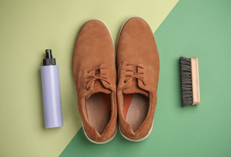 Brown suede shoes are placed on a two-tone green background, flanked by a purple spray bottle on the left and a wooden brush on the right.