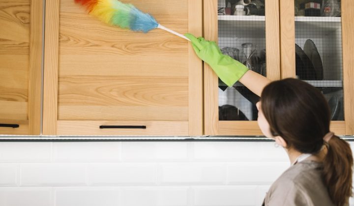 A woman with curly hair smiles while cleaning a table with a blue cloth. A small potted plant is beside her, creating a homely, cheerful atmosphere.