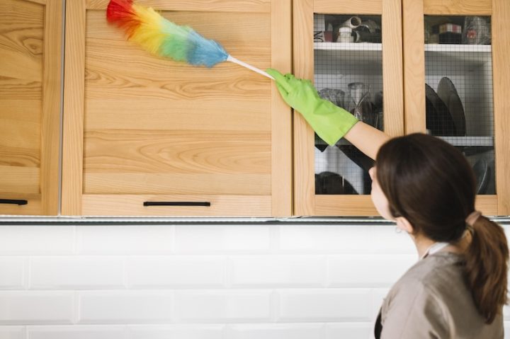 A woman with curly hair smiles while cleaning a table with a blue cloth. A small potted plant is beside her, creating a homely, cheerful atmosphere.