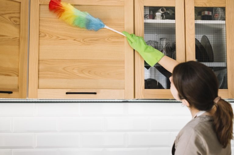 A woman with curly hair smiles while cleaning a table with a blue cloth. A small potted plant is beside her, creating a homely, cheerful atmosphere.