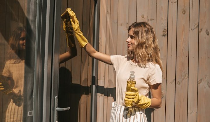 A woman wearing yellow gloves cleans a glass door with a cloth and spray bottle. Sunlight illuminates the wooden wall behind her, creating a warm, focused scene.