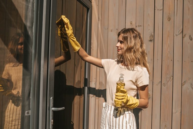 A woman wearing yellow gloves cleans a glass door with a cloth and spray bottle. Sunlight illuminates the wooden wall behind her, creating a warm, focused scene.