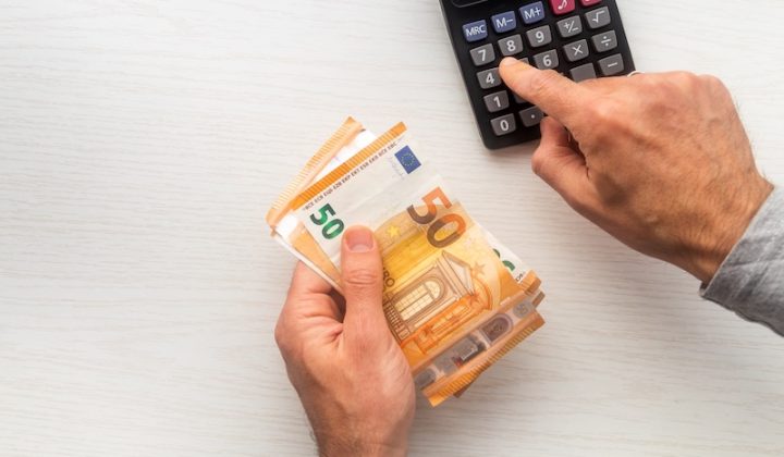 A person holds several 50 euro banknotes in one hand while using the other to press buttons on a calculator, suggesting financial calculation.