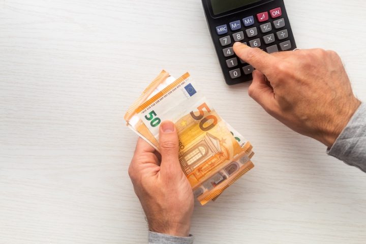 A person holds several 50 euro banknotes in one hand while using the other to press buttons on a calculator, suggesting financial calculation.