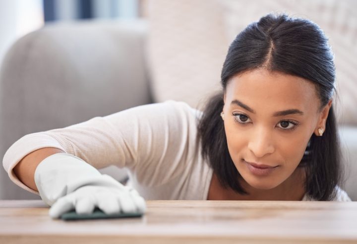 A focused woman cleans a wooden surface with a cloth, wearing a white shirt and gloves. Her expression shows concentration and attention to detail.